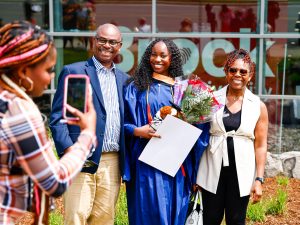 A Brock University graduate poses for a photo outside in her gown surrounded by loved ones. She is holding a bouquet of flowers. Another loved one takes their photo with a smartphone.