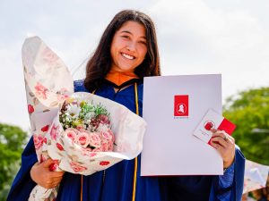 A Brock University graduate poses for a photo outside in her gown while holding a bouquet of flowers and a diploma.