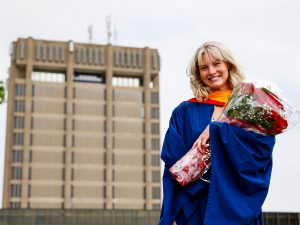 A new Brock University graduate smiles for a photo while holding a bouquet of flowers. The Schmon Tower is visible behind her.