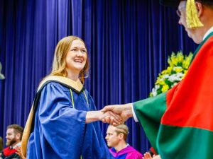 A woman in a graduation gown shakes the hand of a faculty member on stage during Convocation.