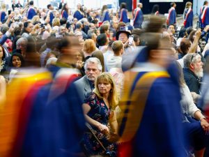 Blurred individuals wearing convocation robes walk in front of an in-focus crowd of seated people who are looking on.