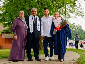 A person in a graduation gown poses with three loved ones while holding her diploma and a bouquet of flowers.