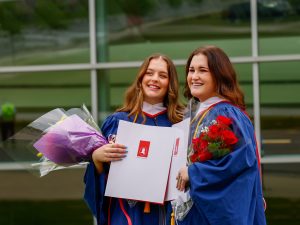 Two women in convocation robes who are holding flowers and their university degrees stand having their photo taken in front of a glass wall.