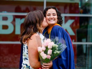 A woman in a convocation robe smiles while holding flowers and receiving a kiss on the cheek from another woman.