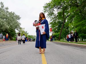 A woman in a convocation robe stands smiling in the middle of a road while holding a stuffed bear and a university degree.