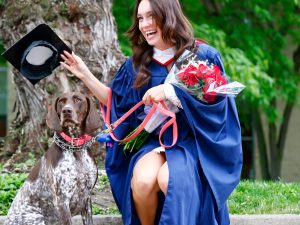A smiling woman in a convocation robe holds flowers while kneeling to put a flat convocation hat on her dog.
