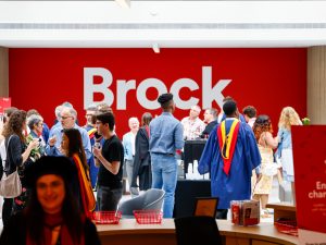 A large group of people, many of whom are wearing convocation robes, mingle in an indoor atrium that has a red wall with a Brock University logo prominently displayed.