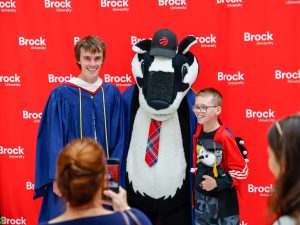 A badger mascot wearing a convocation robe stands between a man wearing a convocation robe and a young boy holding a stuffed animal, all of whom are standing in front of a red backdrop displaying the Brock University logo.