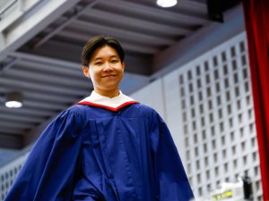 A student in a convocation robe smiles while standing in an indoor gymnasium.