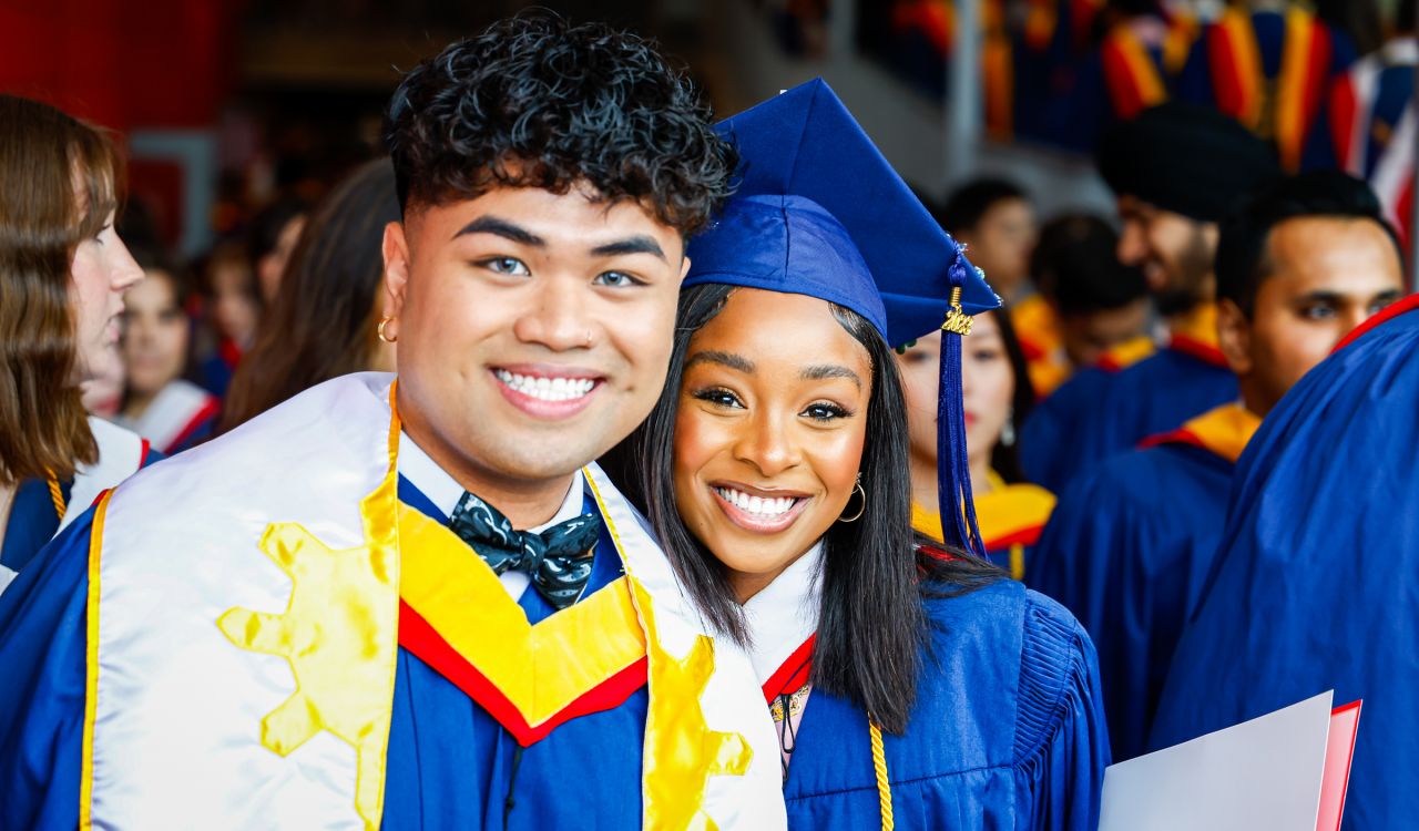 Two graduates in their ceremonial gowns stand in a crowd of graduates.