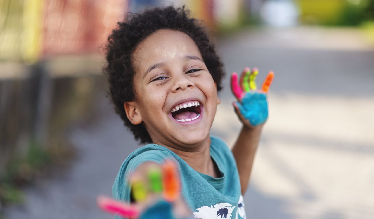A kindergarten student plays with paint outside. He is smiling and showing off his paint-covered hands.
