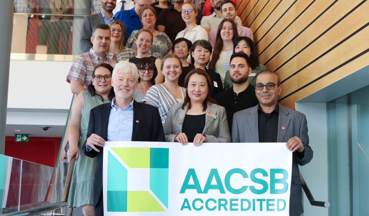 A large staircase is filled with people. At the bottom of the staircase, three people hold a sign stretching the width of the staircase that says AACSB Accredited and has a green square logo.