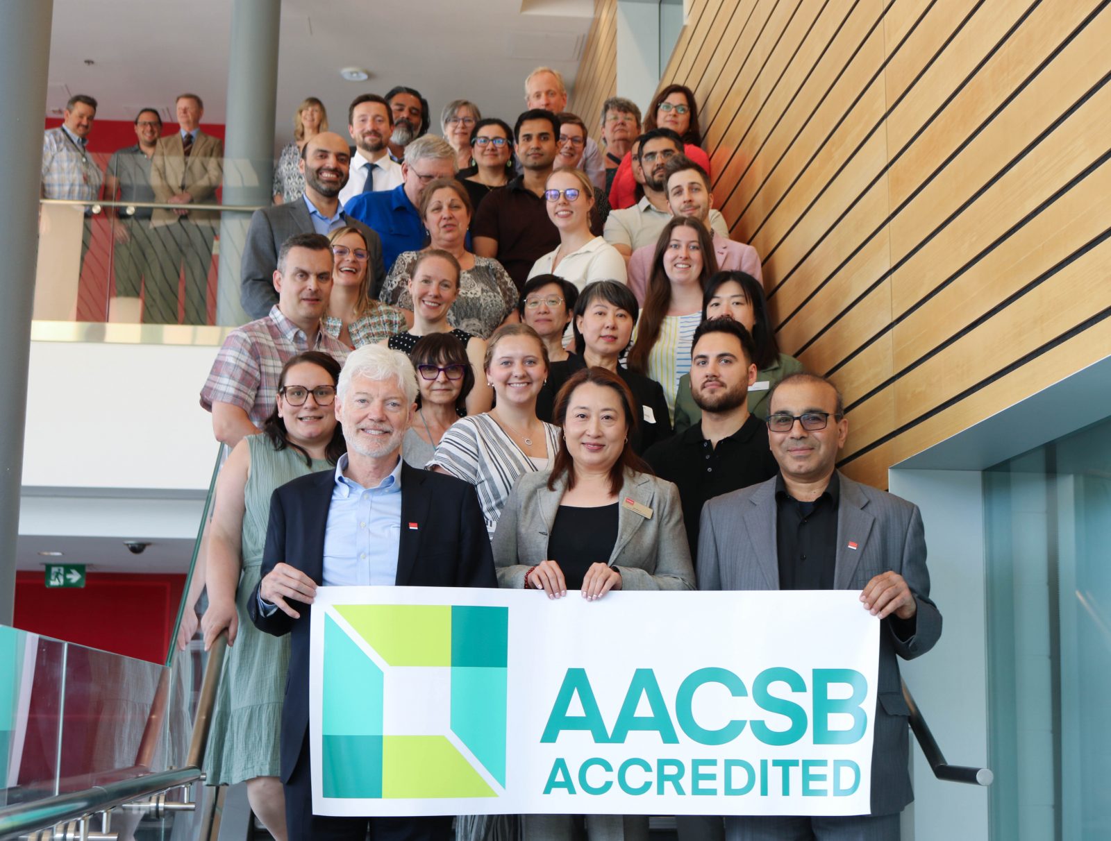 A large staircase is filled with people. At the bottom of the staircase, three people hold a sign stretching the width of the staircase that says AACSB Accredited and has a green square logo.