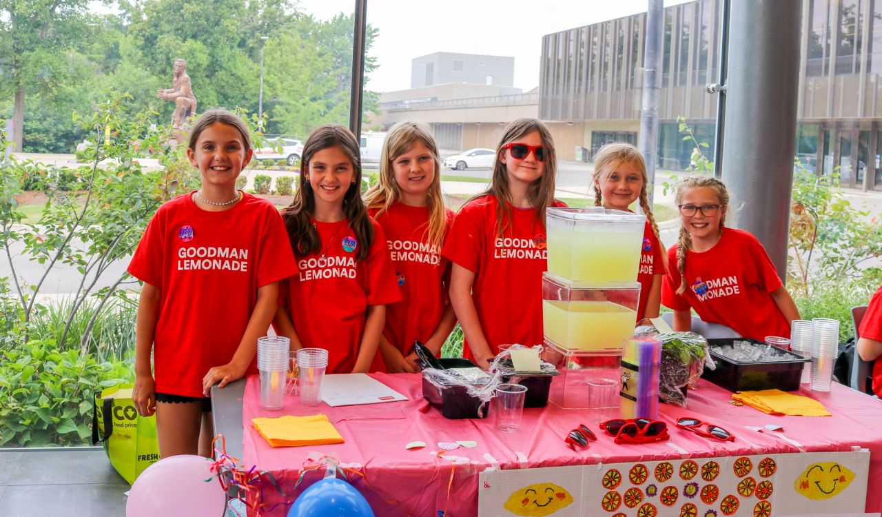 Six elementary school students stand in a line behind a lemonade stand.