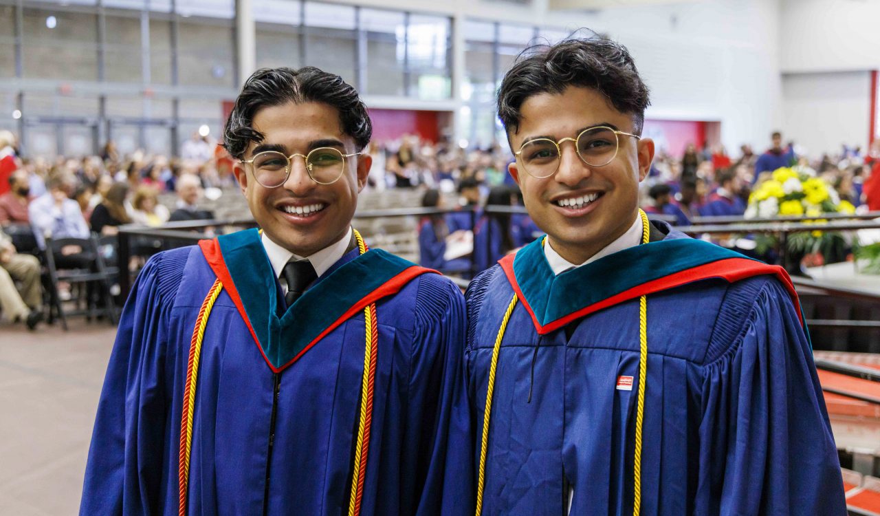 Two graduates stand together in a gymnasium.