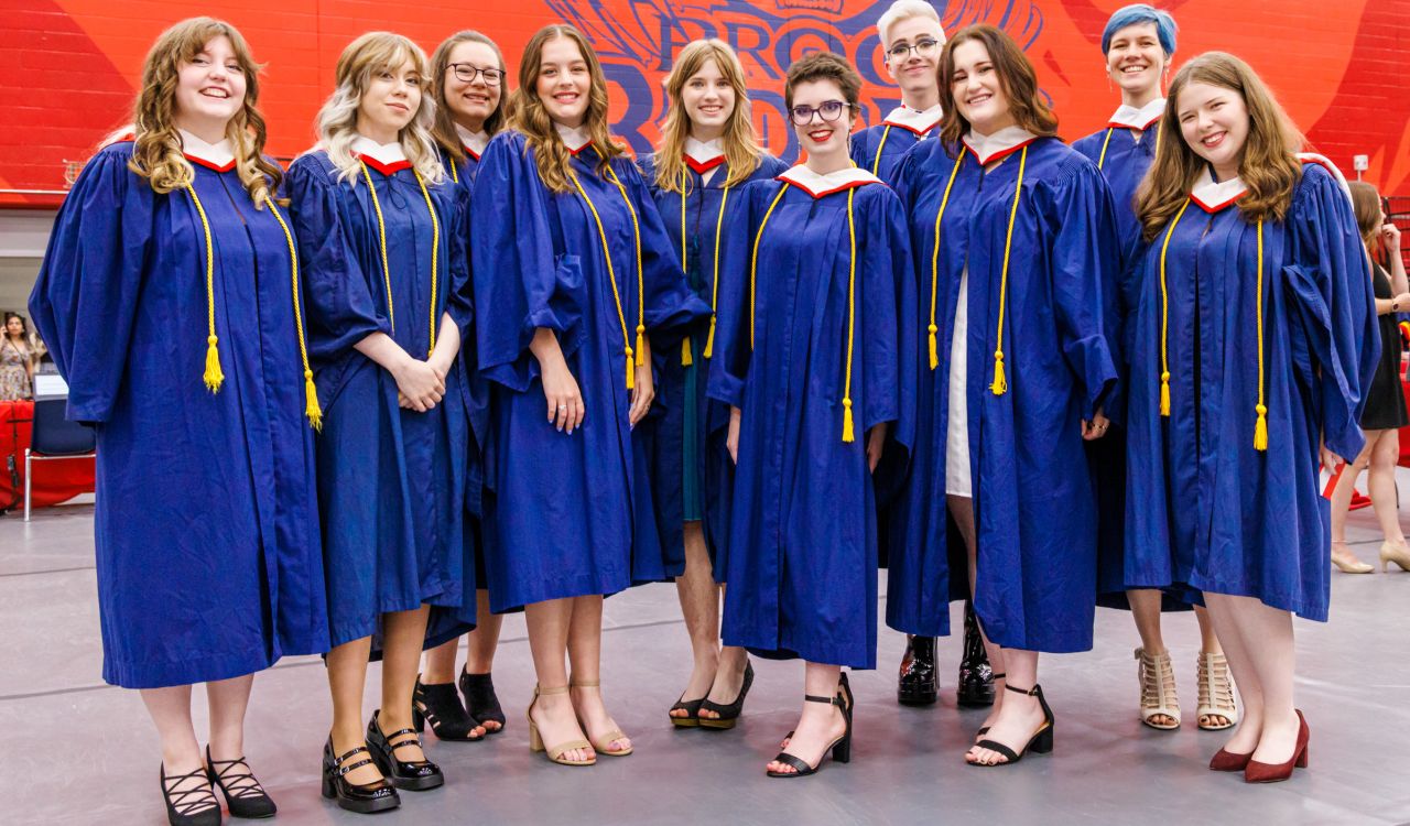 A group of smiling graduates stand together in a university gymnasium with a red wall painted behind them showing an image of the Brock badger. The beaming graduates are wearing bright blue robes with white and red hoods.
