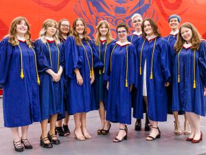 A group of smiling graduates stand together in a university gymnasium with a red wall painted behind them showing an image of the Brock badger. The beaming graduates are wearing bright blue robes with white and red hoods.