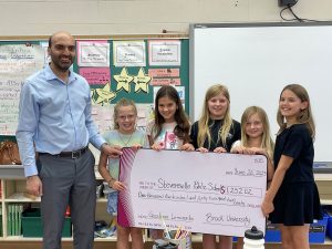 A man hands a novelty cheque to a group of five elementary school students in a classroom.