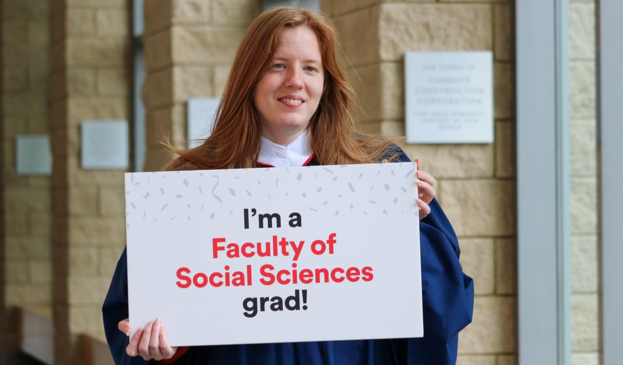 Karli Woods wears a Brock Convocation gown while holding a sign that reads, “I’m a Faculty of Social Sciences grad!”