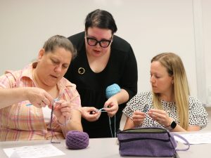 Two women are seated while holding a crochet needle and yarn. A third women, also holding a crochet needle and yarn, stands between them to provide instruction.