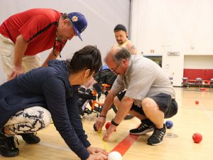 – A man and a woman are kneeling over a measuring tape that is laid across a gym floor between two bocci balls. Another man stands over them while a man in a wheelchair looks down at the measurements.