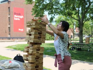 In a park, a woman places a wooden block on top of a large Jenga stack that is on a picnic table.