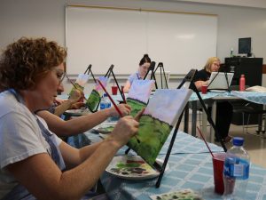 A woman paints on a canvas in a classroom while other students work on their paintings in the background.