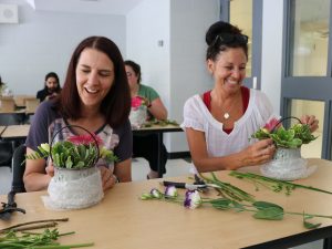 Two woman sit at a table in a classroom each placing cut flowers into ceramic planters.