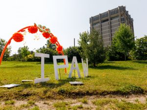 A crescent of orange decorations sits above the letters IPAW in a field in front of a large concrete building.