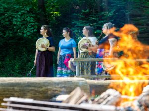 Four women stand holding drums behind a burning fire pit.