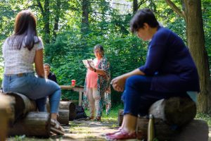 A woman stands reading from a book to a crowd of people sitting on logs in an outdoor setting.