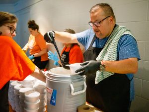 A man serves soup next to women in orange shirts who are preparing other food items.