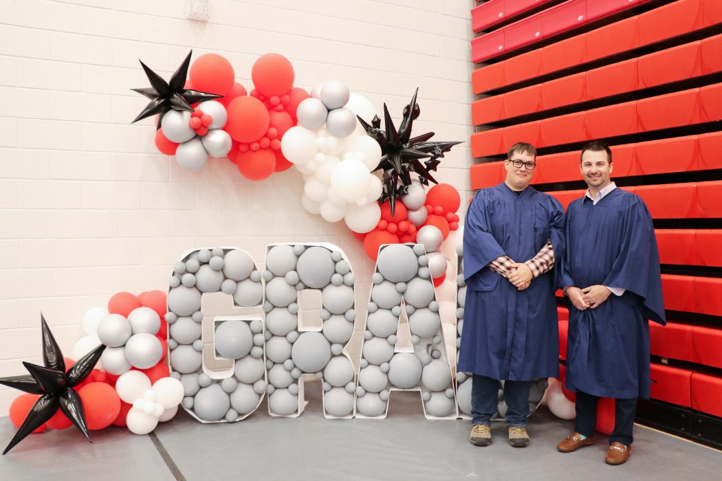 Two graduates in blue Convocation robes stand in front of a balloon display that spells out the word 'GRAD.'