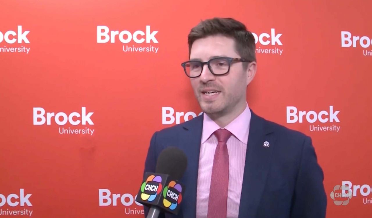 A man in a suit speaks into a microphone in front of a red backdrop that says 'Brock University'