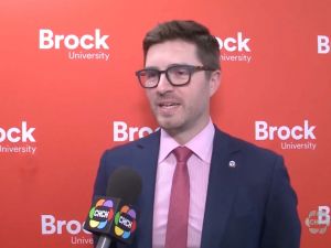 A man in a suit speaks into a microphone in front of a red backdrop that says 'Brock University'