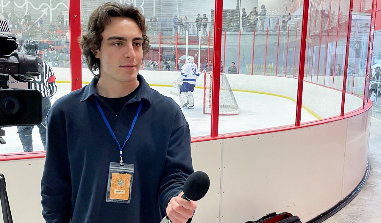 Brock University student Lucas Rotondo stands next to a video camera and holds a microphone in his hand. Behind him is an ice rink with hockey players.
