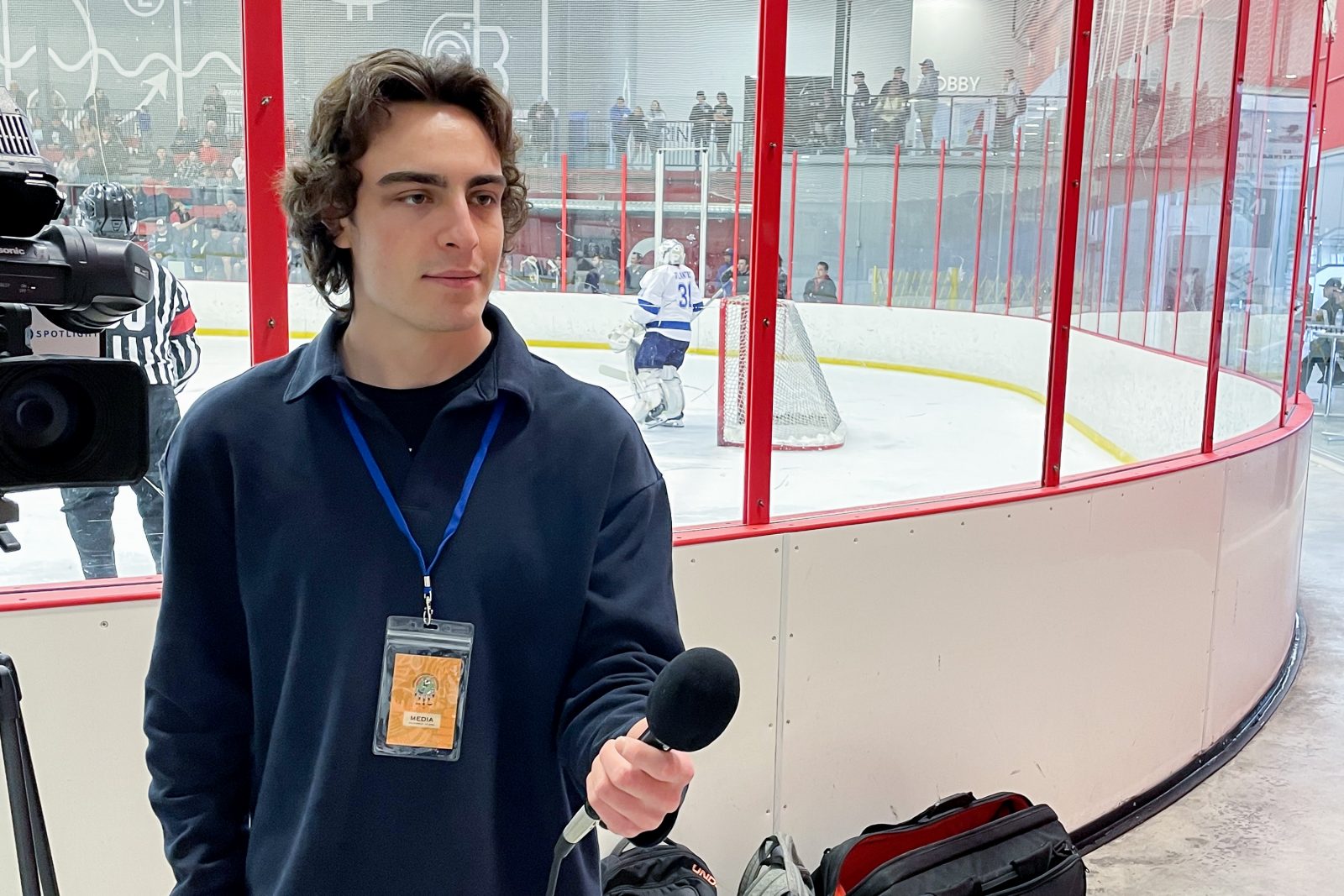 Brock University student Lucas Rotondo stands next to a video camera and holds a microphone in his hand. Behind him is an ice rink with hockey players.