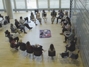 Viewed from above, about 20 seated participants face each other for discussion in a brightly lit room.