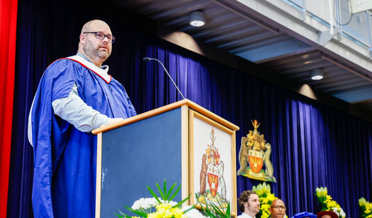 A man in an academic gown speaks from behind a podium on stage during Convocation.