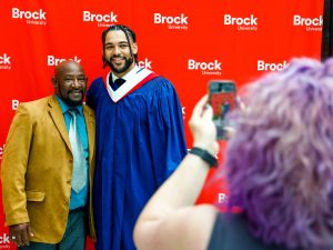 A new graduate poses for a photo with a loved while someone takes their picture using a smartphone.