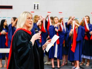 A woman in an academic gown speaks to a group of graduating university students wearing graduation gowns.