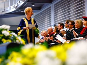 A woman walks across the stage carrying documents during a university graducation ceremony.