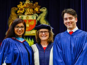 Three people in academic gowns stand side-by-side on stage during a Brock University graduation ceremony. The University's crest is visible in the background.