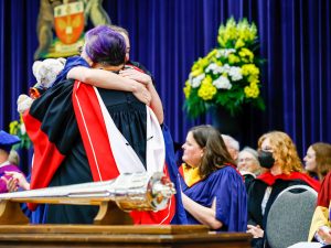 A faculty member hugs a graduating student on stage during a university graduation.
