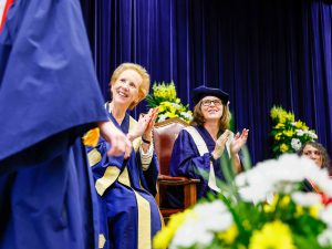 Two women on stage applaud during a university graduation ceremony.