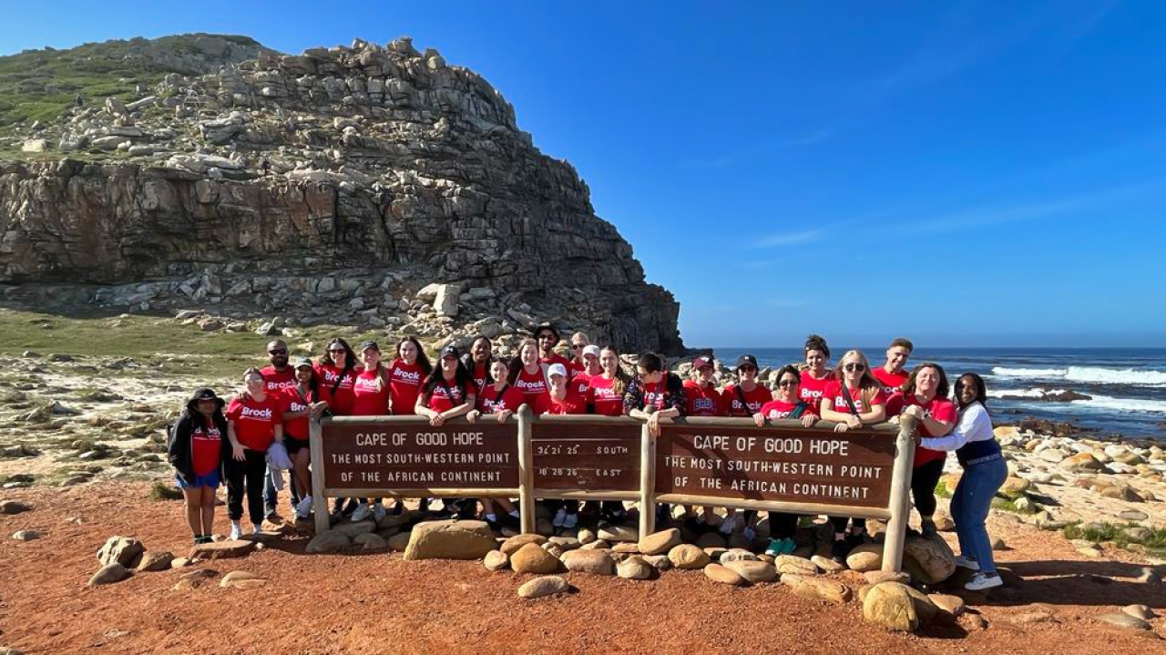 A large group of students wearing red Brock T-shirts stand behind signs that read “Cape of Good Hope: The most south-western point of the African continent.” Behind them is a large, rocky peninsula and a vast body of water.