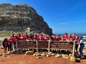 A large group of students wearing red Brock T-shirts stand behind signs that read “Cape of Good Hope: The most south-western point of the African continent.” Behind them is a large, rocky peninsula and a vast body of water.