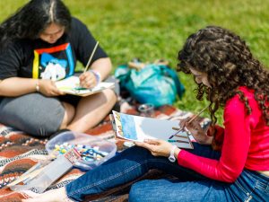 Two women sit on a blanket in a grassy field while painting on pieces of paper.