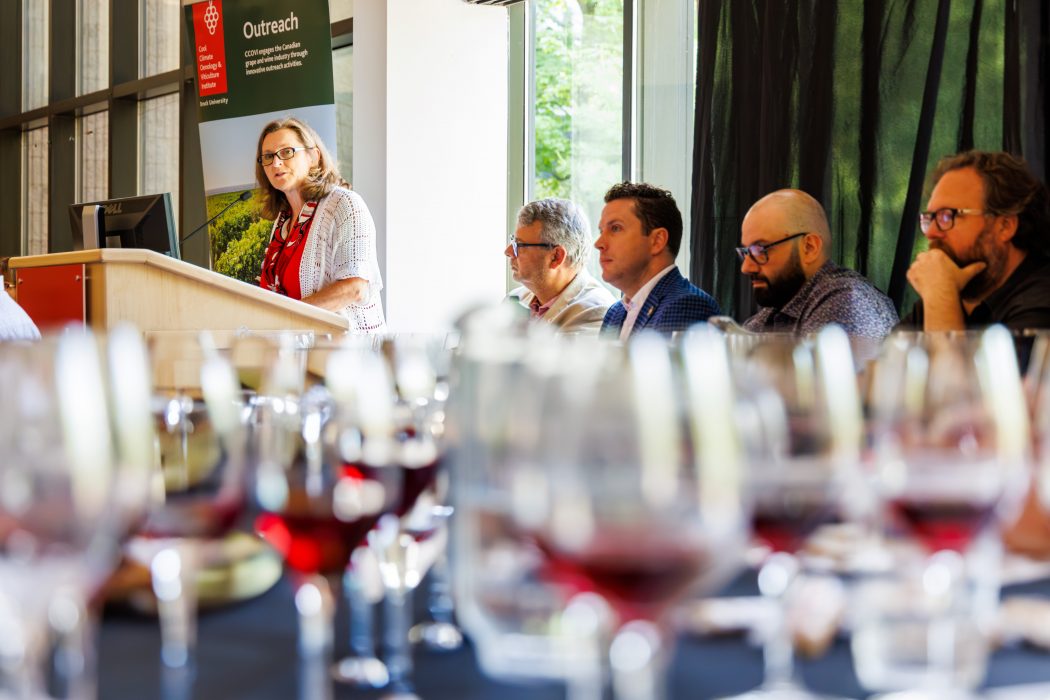 A woman speaks at a podium with a group of four people sitting at a table beside her. In the foreground are out-of-focus glasses of red wine.
