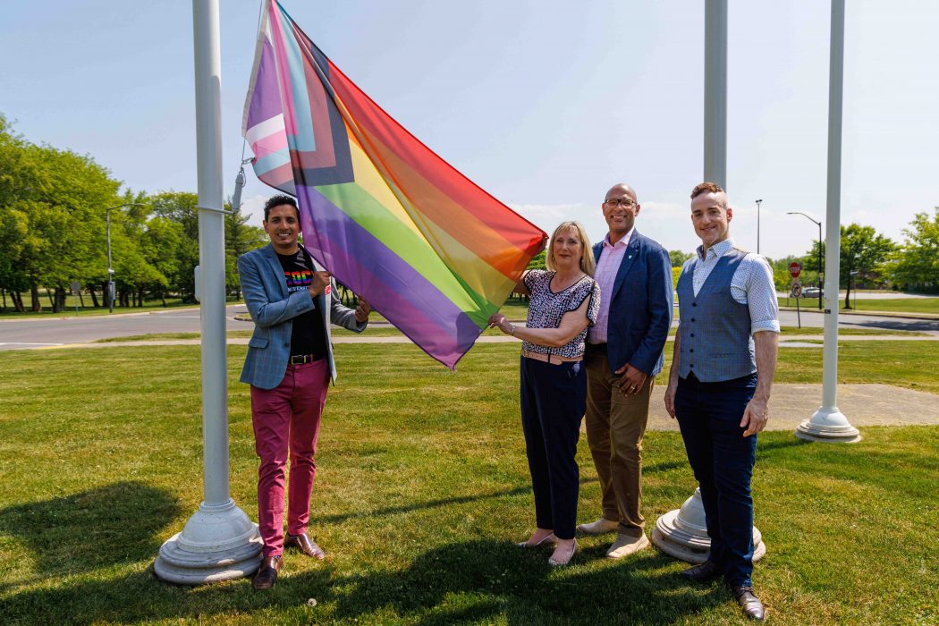 Four people stand holding a multicoloured flag with chevrons on the end as it is prepared to be raised up a flag pole in front of Brock University.